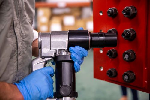 The image shows a close-up view of a person using a shear wrench on tension control (TC) bolts. The person, whose face is not visible, is wearing blue gloves and is working on a red structure with multiple black bolts. This setting suggests a focus on precision and safety in a construction or manufacturing environment. The wrench itself is metallic and robust, designed for heavy-duty tasks.